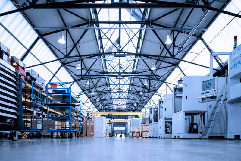 Spacious modern factory interior with high ceilings, industrial lighting, machines, equipment, and storage shelves lining both sides of a wide central aisle.