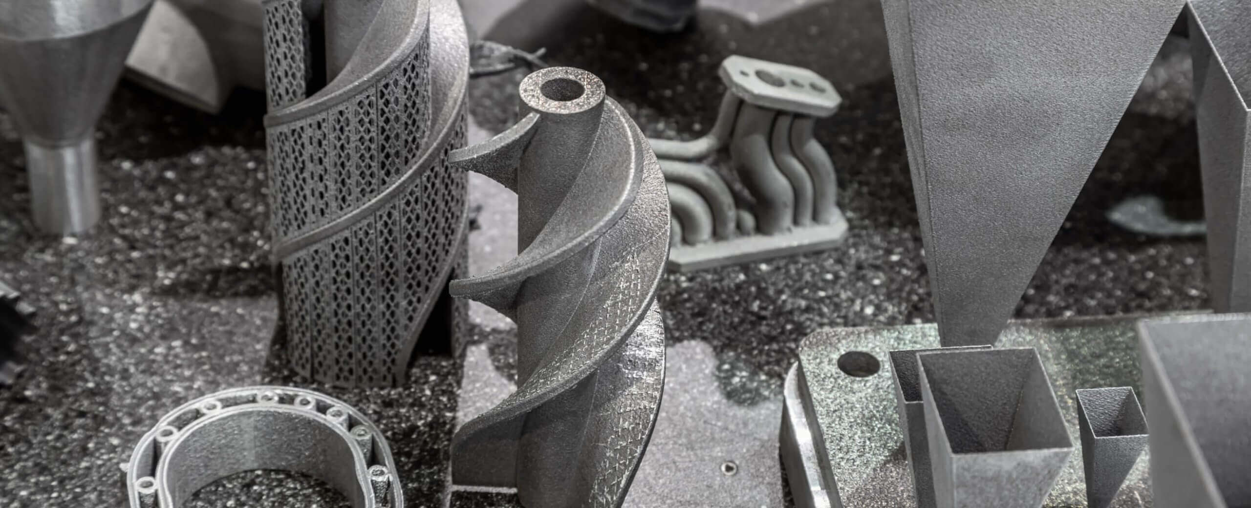 A close-up view of various intricate metal parts and components, likely produced using 3D printing or additive manufacturing, displayed on a textured metallic surface.