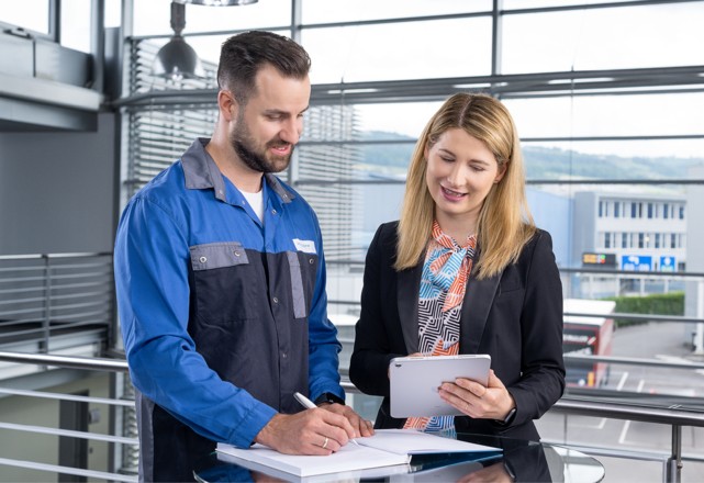 An employee in work clothes and a businesswoman are reviewing documents on a tablet in a modern office building