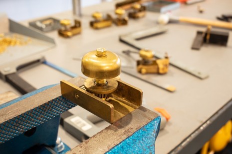 Eine Klingel ist in einem Schraubstock eingespannt. Dahinter sieht man auf der Werkbank Werkzeug liegen. 