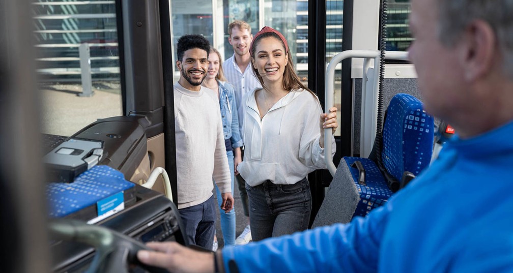 Zwei junge Frauen und Männer steigen in den Stahlwelt Werkstour Bus ein und lächeln. Der Busfahrer begrüßt sie. 