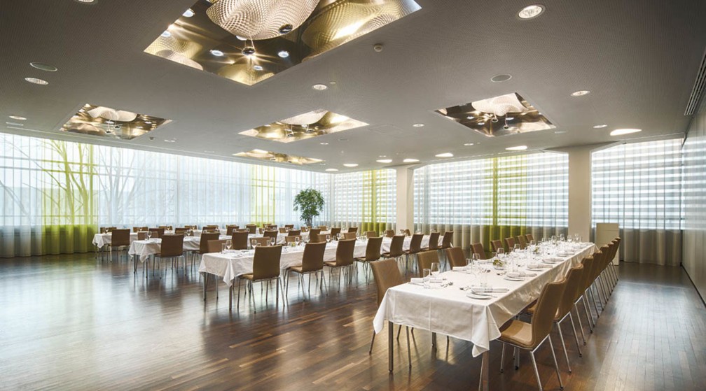 Ein heller Veranstaltungssaal mit vier Reihen gedeckten Tischen und Stühlen. 