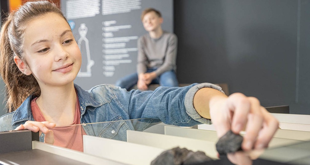 Ein Mädchen nimmt ein Stück Erz aus einer Lade, ein Junge schaut ihr zu. 