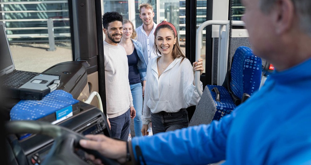 Zwei junge Frauen und Männer steigen in den Stahlwelt Werkstour Bus ein und lächeln. Der Busfahrer begrüßt sie. 
