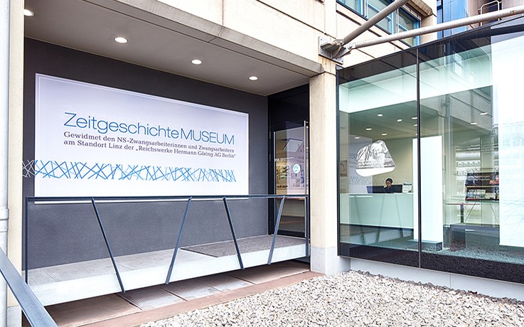 Eingang des ZeitgeschichteMUSEUMs. Links neben dem Eingang befindet sich ein weißes Schild mit dem Titel des Museums "ZeitgeschichteMUSEUM". Rechts neben dem Eingang ist eine Glasfront. 